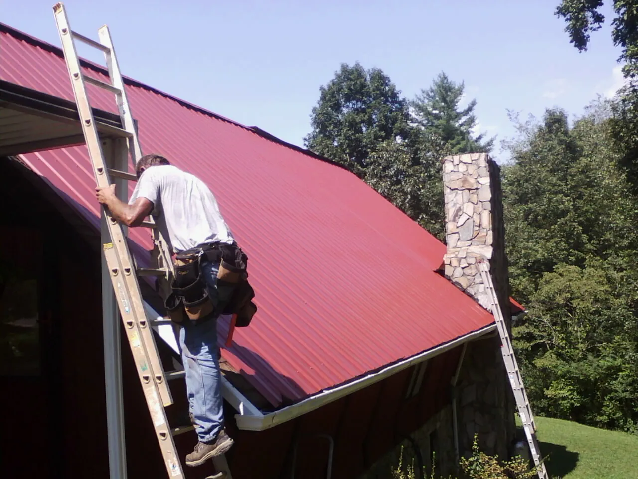 Roofer on a ladder beside a red metal roof and stone chimney — Soffit & Fascia Repair by Roofing Brothers in Kanosh
